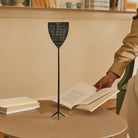 Een persoon houdt een open boek vast aan een ronde tafel, waar een ander boek en de Alessi Dr. Skud Vliegenmepper Grijs van Alessi staan. Boeken vullen de planken op de achtergrond.