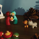 Twee levendige Alessi Happy Eternity babyfiguren tonen Maria en Jozef met baby Jezus in een kerststal, omringd door een ezel, os, dennenappels en kerstornamenten op een warm verlicht houten oppervlak.