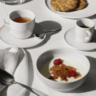 Een witte kom yoghurt met frambozen en een koekje, naast een lepel, thee, een espresso geserveerd in de Alessi Itsumo Koffiekop + Schotel Wit 21 cl, koekjes op een bord en een servet - alles op een wit tafelkleed geïnspireerd op Japans design.