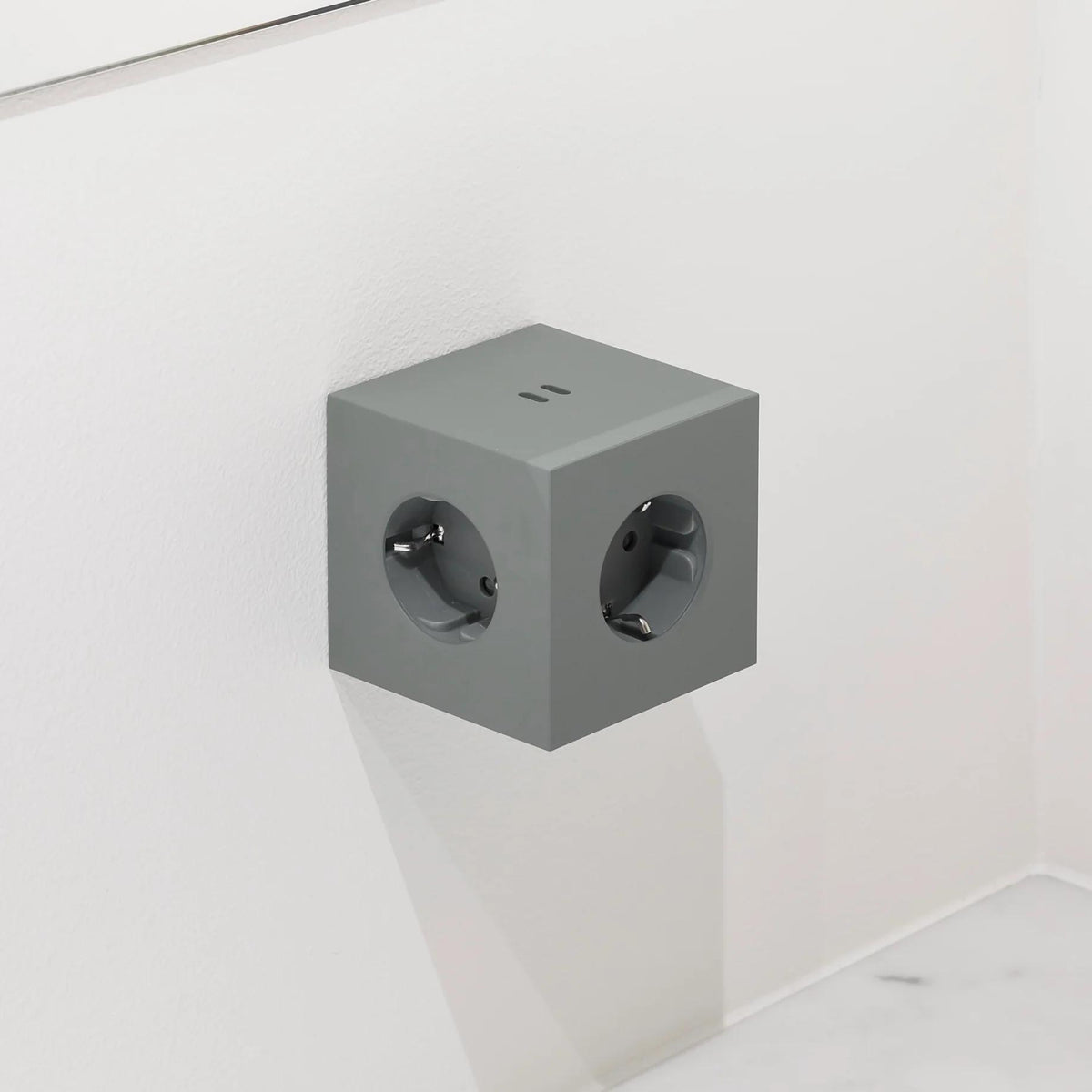 Een Avolt Square 2 USB-C Stekkerdoos Eik Groen, een kubusvormige stekkerdoos met meerdere stopcontacten en USB-C uitbreiding, van het merk Avolt, is gemonteerd op een witte muur boven een wit oppervlak.