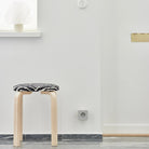 Een minimalistische kamer met een licht houten krukje met een zwart-witte zitting, een grijze vloer, een witte muur met een Avolt Square 2 USB-C Stekkerdoos Gotland Grijs van Avolt en een vensterbank met een wit decoratief object.