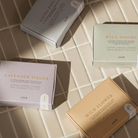 Vier Azur Lavender Fields Natuurlijke Zeep Bar dozen rusten op bleke tegels in het zonlicht, hun zachte kleuren hinten naar aromatherapie van Azur voor een ultieme ontspannende douche-ervaring.