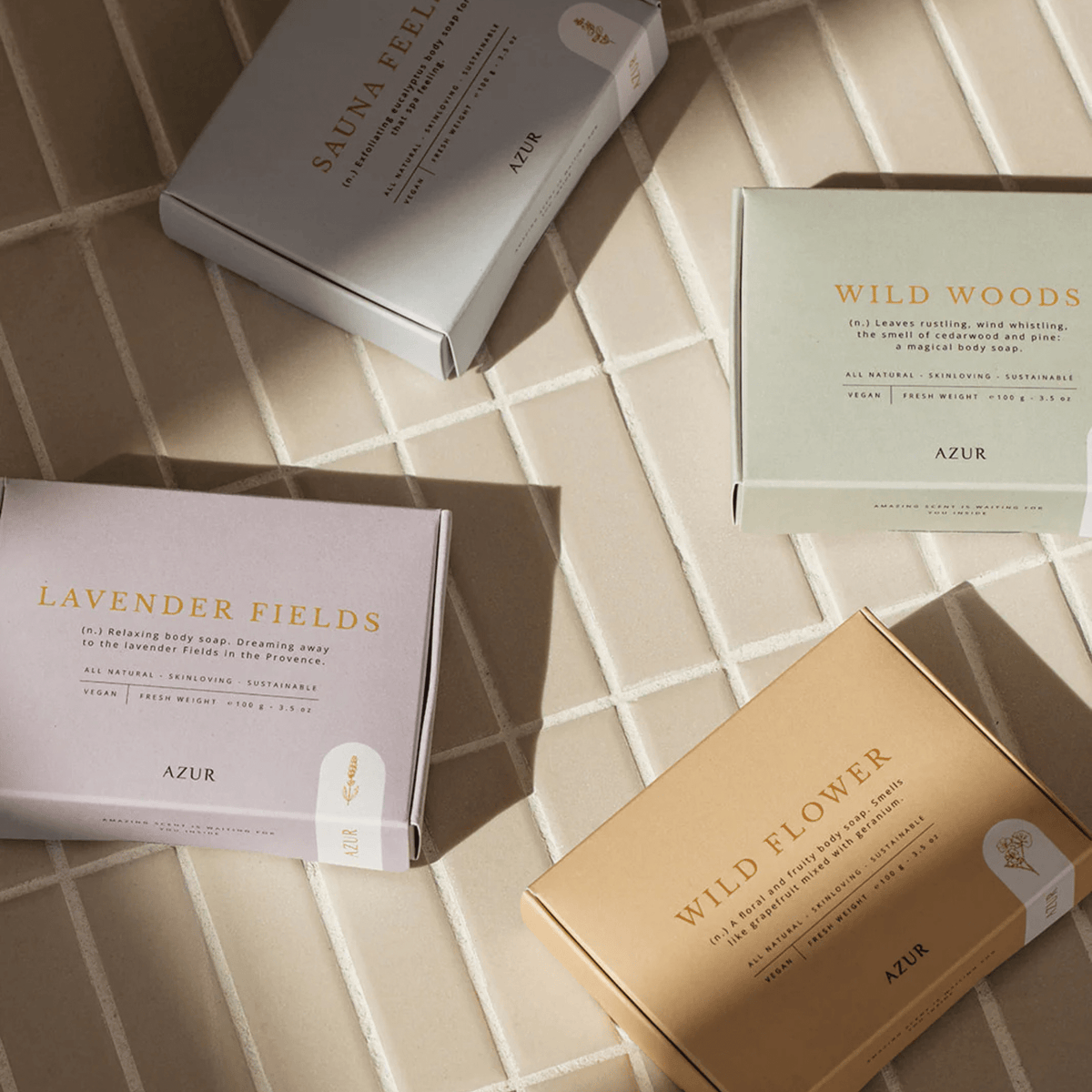 Vier Azur Lavender Fields Natuurlijke Zeep Bar dozen rusten op bleke tegels in het zonlicht, hun zachte kleuren hinten naar aromatherapie van Azur voor een ultieme ontspannende douche-ervaring.