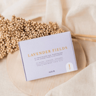 Een lavendelblauwe Azur Lavender Fields Natuurlijke Zeep Bar zit op een licht rond oppervlak met een tros gedroogde bloemen ernaast - perfect voor een ontspannende douche - tegen een zacht beige stof op de achtergrond.