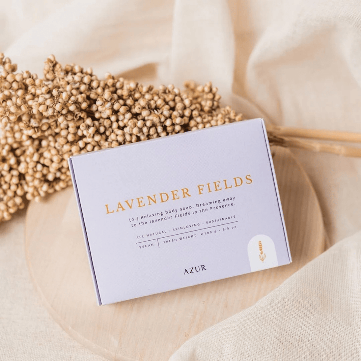 Een lavendelblauwe Azur Lavender Fields Natuurlijke Zeep Bar zit op een licht rond oppervlak met een tros gedroogde bloemen ernaast - perfect voor een ontspannende douche - tegen een zacht beige stof op de achtergrond.
