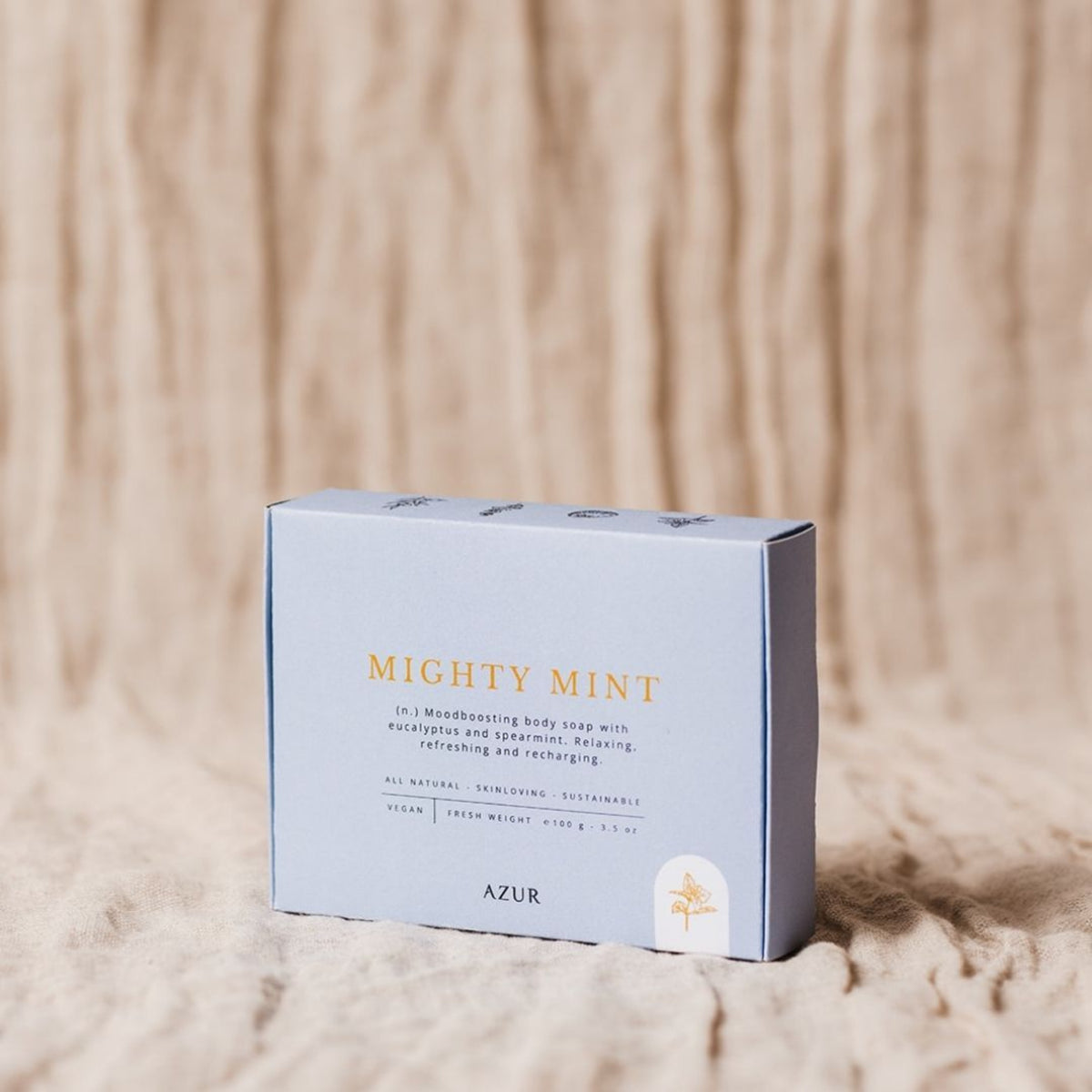 Een lichtblauwe doos Azur Mighty Mint Natuurlijke Zeep Bar rust op beige structuurstof. De duurzame verpakking benadrukt de verfrissende, ontspannende en veganistische eigenschappen met gouden en witte tekst op de voorkant.