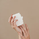 Een persoon met netjes gemanicuurde nagels houdt de Azur Sauna Feels Natuurlijke Scrub Zeep Bar vast tegen een effen beige achtergrond, die de eenvoud van Azur's natuurlijke zeep benadrukt.