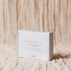Een witte doos Azur Sauna Feels Natuurlijke Scrub Zeep Bar rust op beige structuurstof. Deze duurzame, volledig natuurlijke, veganistische en ongeparfumeerde zeep is perfect voor zachte dagelijkse verzorging als milde scrubzeep.