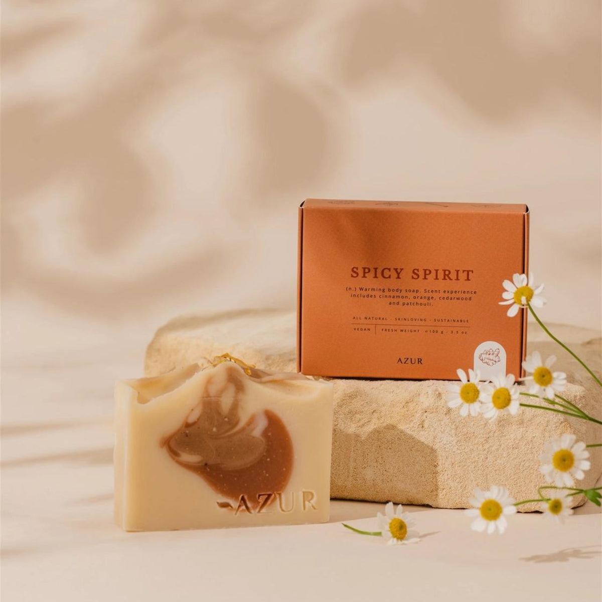 De Azur Spicy Spirit Natuurlijke Zeep Bar rust op beige steen naast zijn oranje doosje. Witte en gele madeliefjes staan op de voorgrond tegen een zachte, neutrale achtergrond. Geïnspireerd door kurkuma.