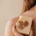 Een persoon met lang haar houdt de Azur Spicy Spirit Natuurlijke Zeep Bar van Azur, met een beige en bruine werveling van kurkuma, tegen zijn zeepschouder. Alleen een deel van de schouder, rug en ketting zijn zichtbaar.