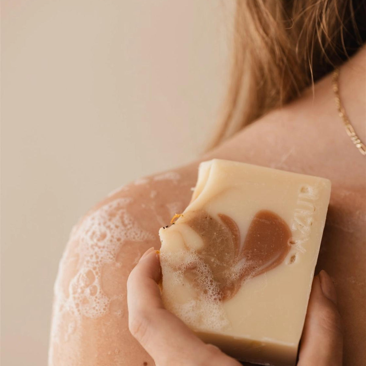 Een persoon met lang haar houdt de Azur Spicy Spirit Natuurlijke Zeep Bar van Azur, met een beige en bruine werveling van kurkuma, tegen zijn zeepschouder. Alleen een deel van de schouder, rug en ketting zijn zichtbaar.