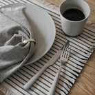 Een keramisch bord met een gestreept servet en een Broste Copenhagen mok met zwarte koffie, plus vork en mes, rusten op een tafeldecoratie placemat met franjes, geaccentueerd door de Broste Ring Servetring 6 st. van Broste Copenhagen op een houten tafel.