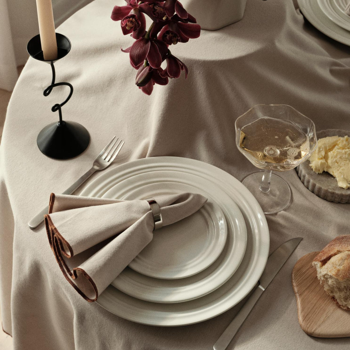 Een beige tafelkleed dekt een ronde tafel gedekt met Broste Copenhagen borden, een Broste Ring Servetring 6 st. met een gevouwen servet, keukengerei, champagneglas, brood, kandelaar, boterbordje en een vaas met donkerpaarse orchideeën voor een elegante tafeldecoratie.
