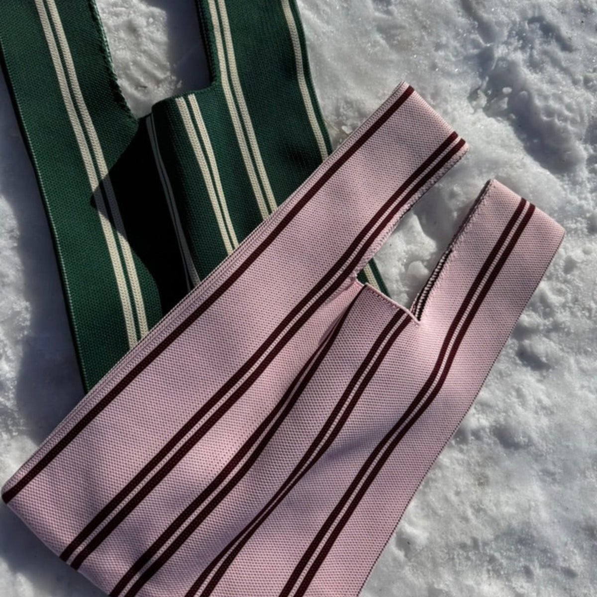 Twee Caro Antwerp Handtas Double Dash 2.0 Roland Larose tote bags-één groen/wit, één roze/bordeaux met verticale strepen-liggen op sneeuw in fel zonlicht. Deze stijlvolle geweven tassen van Caro Antwerp zijn perfect voor dagelijks gebruik.