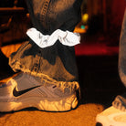 Een persoon in een gerafelde blauwe spijkerbroek en witte Nike sneakers draagt een Caro Antwerp Caro The Scrunch Club Scrunchie 2 st. om één broekspijp. De schemerige scène heeft een warme, stemmige sfeer.
