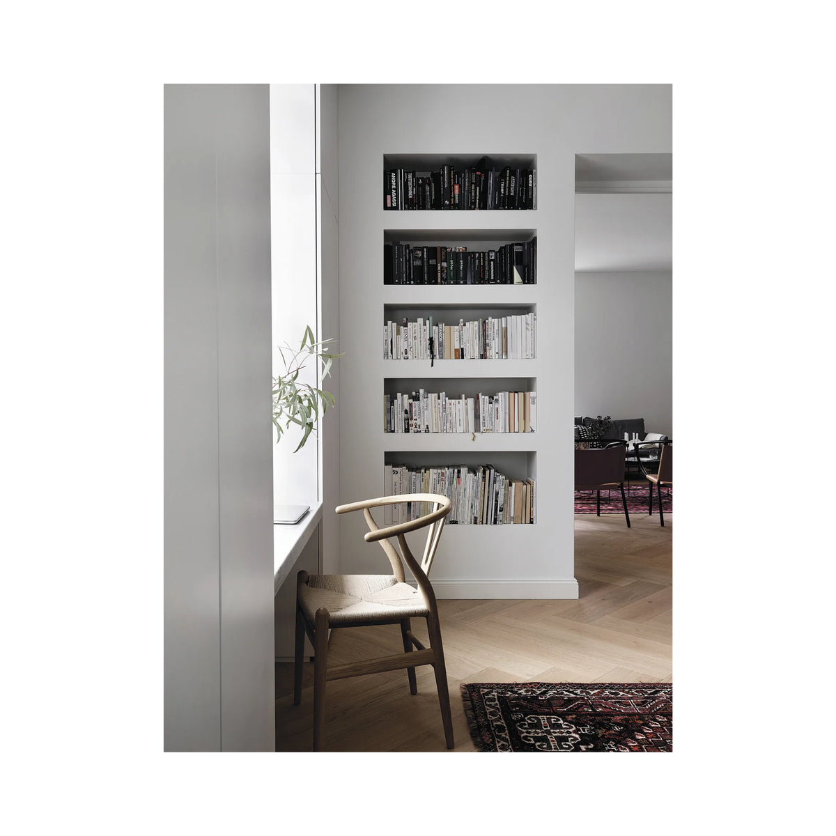 Een minimale boekenplank ingebouwd in een witte muur toont netjes gerangschikte boeken, waaronder het Cozy Publishing Happy Homes Creative Boek. Een houten stoel staat bij het raam, met een glimp van Scandinavisch geïnspireerde eetkamermeubels op de achtergrond.