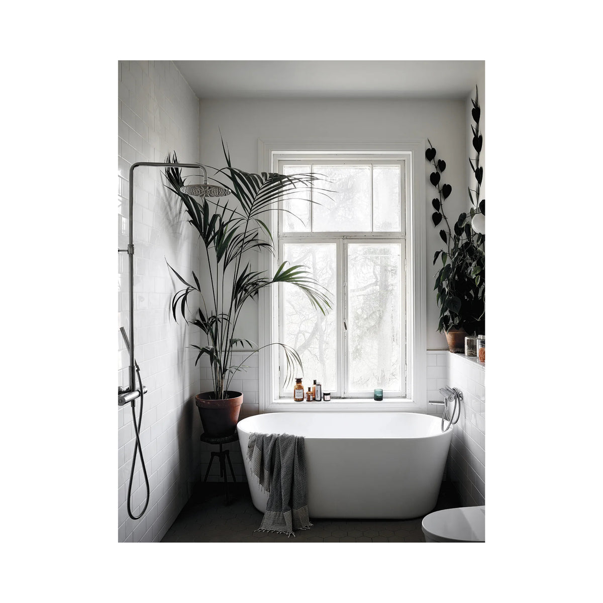 Een moderne badkamer met een wit vrijstaand bad, planten in potten en flessen op de vensterbank toont Scandinavisch design uit het Cozy Publishing Happy Homes Creative Boek, terwijl zonlicht de ruimte vult en een handdoek op het bad rust.