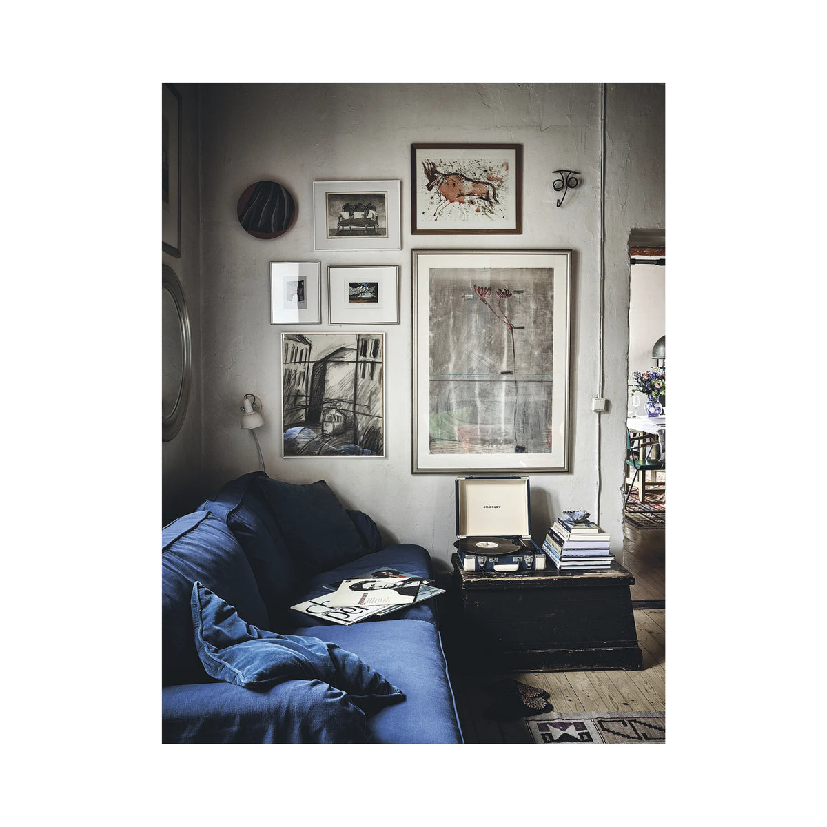 Een gezellige woonkamerhoek met een blauwe bank, ingelijste kunst, het Cozy Publishing Happy Homes Creative Boek, een platenspeler en een tafeltje. De ontspannen ruimte straalt vintage charme uit terwijl het zonlicht zachtjes naar binnen valt.