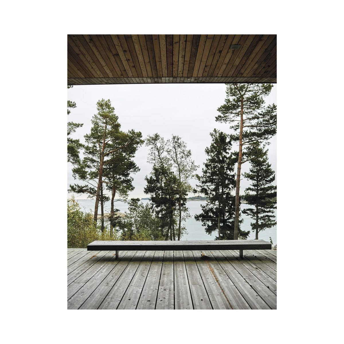 Een minimalistisch houten bankje staat op een ruim terras met uitzicht op pijnbomen en rustig water, omlijst door een modern dak - een uitnodigende scène rechtstreeks uit het Cozy Publishing Happy Homes Hideaways Boek.