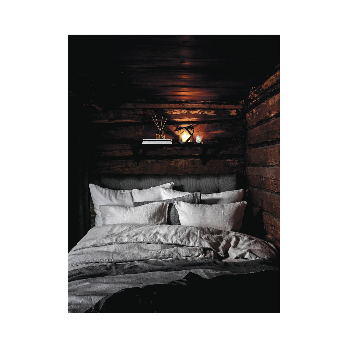 Een knus bed met wit en grijs beddengoed staat tegen rustieke houten muren en weerspiegelt de charme van Finse zomerhuizen. Op een plank daarboven ligt het Cozy Publishing Happy Homes Hideaways Boek, dat bijdraagt aan de uitnodigende sfeer onder een zacht gloeiende lamp.