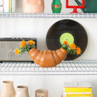 De Fluid Market Croissant Vaas van Fluid Market, een vaas van keramiek in de vorm van een croissant, toont oranje bloemen op een draadplank met kleurrijke decoraties, vinylplaten, luidsprekers en boeken - ideaal voor een speels interieur met Franse invloeden.