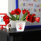 Een Fluid Market Ramen Pot in wit van Fluid Market houdt rode tulpen op een zwarte schouw, naast een maneki-neko kattenbeeldje en ingelijste levendige kunst met speelse tekst-een levendige display voor fans van design en keramieken objecten.