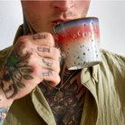 Een getatoeëerd persoon in een groen shirt houdt de HK Living 70s Ceramics Americano Mok Asteroids vlakbij zijn mond, waarbij de gedetailleerde tatoeages op zijn hand en borst goed te zien zijn, evenals een ketting met een ronde hanger.