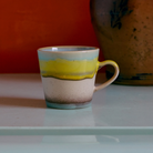 De HK Living 70s Ceramics Americano Mok Glint, met blauwe, gele en bruine golvende strepen, staat op een lichtblauwe ondergrond, met op de achtergrond een grote bruine vaas tegen een oranje muur.