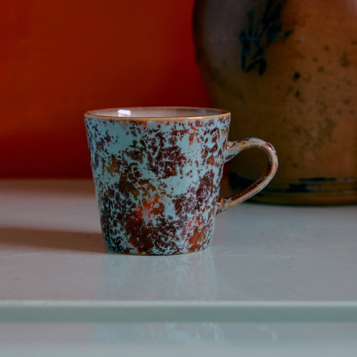De HK Living 70s Ceramics Americano Mok Patina, met een gevlekte blauw-bruine glazuurlaag, staat op een lichtgekleurde ondergrond met daarop een vaag bruin aardewerkvoorwerp en daarachter een oranje achtergrond.