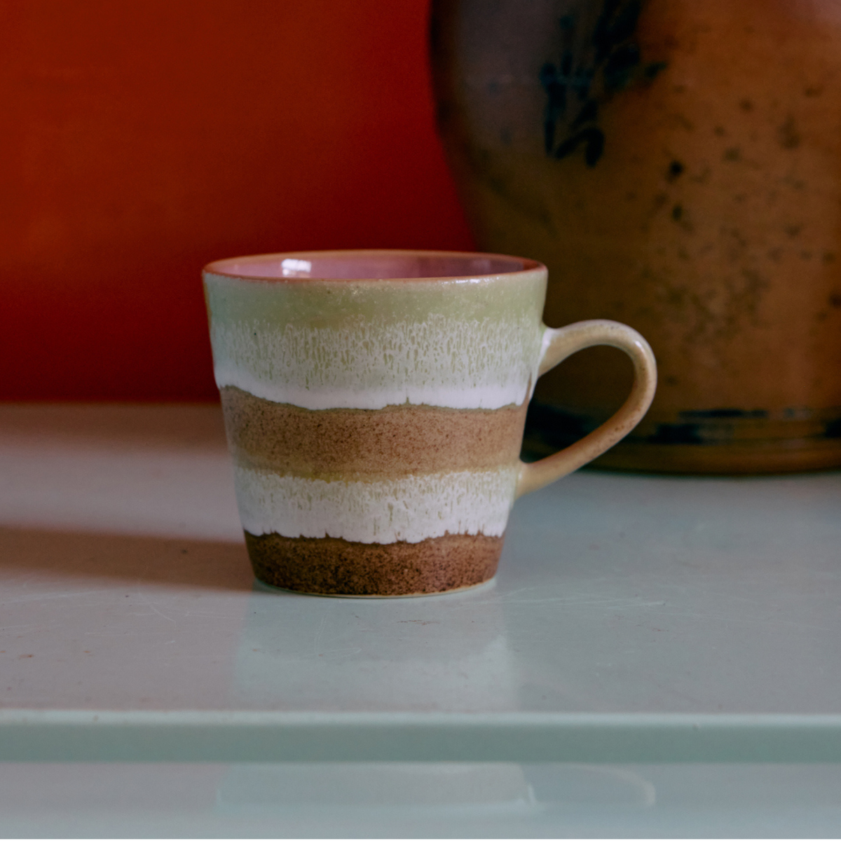 Een HK Living 70s Ceramics Americano Mok Umber, met aardse bruine, beige en witte strepen, staat op een licht oppervlak, met op de achtergrond een grote, wazig afgebeelde bruine kleipot tegen een roodachtige muur.