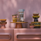 De HK Living 70s Ceramics Americano Mok Sunbeam heeft aardse tinten en patronen met textuur, prachtig uitgestald op een roze dressoir tegen een zachtroze muur met doorvallend zonlicht.