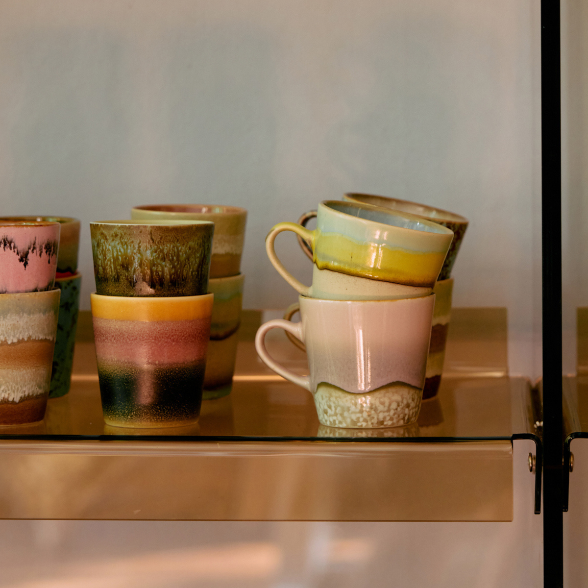 Een reeks kleurrijke keramische mokken, waaronder de HK Living 70s Ceramics Americano Mok Muse met diverse patronen en glazuren, staat opgesteld op een gouden plank tegen een lichte achtergrond.