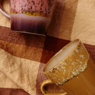 De HK Living 70s Ceramics Americano Mok Sunbeam is voorzien van gevlekte, aardse glazuren in roze, paars, geel en bruin, afgezet tegen een bruin-beige geruite stof — wat de charme van een retro koffiemok oproept.