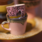 Een close-up van twee op elkaar gestapelde HK Living 70s Ceramics Americano Mok Blast-kopjes, het ene roze en het andere lichtroze met een groenachtige rand, op een geel gespikkeld schoteltje. Perfect voor je Americano. Merk: HK Living.