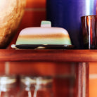 Het HK Living 70s Ceramics Botervlootje Mercury botervlootje staat op een houten plank, met daarachter een grote blauwe vaas en bruin glas, terwijl de glazen potten eronder de retro vibe weerspiegelen tegen een muur van rode bakstenen.