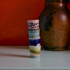 Vier HK Living 70s Ceramics Eierdop Vibrance kopjes zijn gestapeld op een bleek oppervlak voor een aardse kan en oranje wand. Elk kopje heeft een uniek, artistiek glazuur geïnspireerd op de retro eierdop stijl van HK Living.