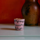 De HK Living 70s Ceramics Koffiekop Bubble, een kleine roze handgemaakte beker met zwarte structuurpatronen, rust op een licht oppervlak, met een grote bruine vaas en een oranje muur op de achtergrond.