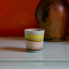 De HK Living 70s Ceramics Koffiekop Glint, met blauwe, gele en bruine strepen, is geplaatst op een licht oppervlak met een grote bruine vaas en een oranje muur op de achtergrond.