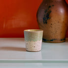 Een kleine HK Living 70s Ceramics Koffiekop Slush met een gespikkeld crème en groen glazuur staat op een licht oppervlak, terwijl een deel van een grote bruine vaas op de achtergrond verschijnt tegen een oranje muur.