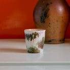 Een HK Living 70s Ceramics Koffiekop Tide, met een aards groen en bruin glazuur, rust op een licht oppervlak. Een grote bruine vaas staat vaag op de achtergrond, tegen een oranje muur.
