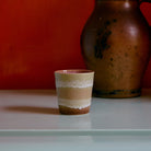 Een kleine HK Living 70s Ceramics Koffiekop Umber, bruin met een crèmekleurig golvend patroon, staat op een licht oppervlak. Daarachter staat een grote bruine aardewerk kan tegen een rood-oranje muur, die de HK Living stijl benadrukt.