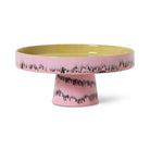 De HK Living 70s Ceramics Kom op Voet Bubble taartstandaard heeft een breed, plat geel oppervlak met een roze glazuur en een zwart druppelpatroon op de rand en basis, wat de kenmerkende jaren 70 stijl van HK Living weerspiegelt.