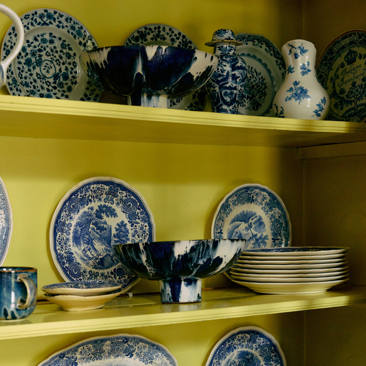 HK Living 70s Ceramics Kom op Voet Indigo van HK Living valt op tussen blauw en wit porseleinen borden, kommen en vazen met traditionele bloem- en landschapsmotieven gerangschikt op gele planken - elk stuk een decoratief statement.