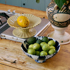 Een keramieken sokkel met een geel gespikkelde vrucht staat bij de HK Living 70s Ceramics Kom op Voet Indigo gevuld met groene limoenen en avocado's op een houten aanrecht naast een grote statement vaas.