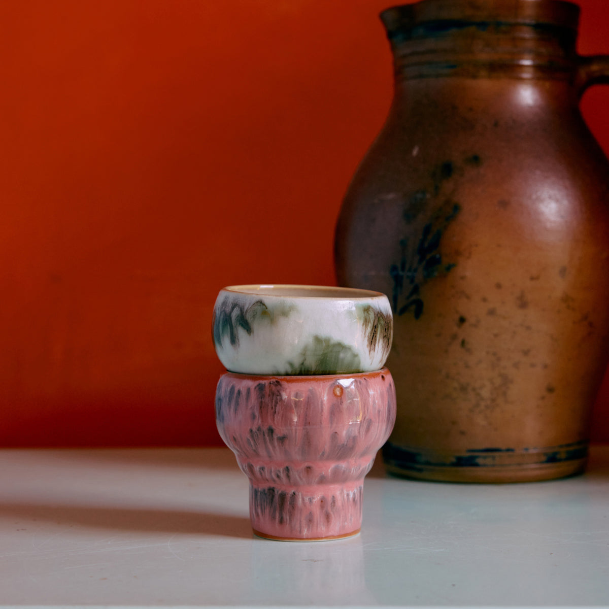 Twee kleine HK Living 70s Ceramics Lungo Mokken Expression-bekers - een met groen en wit patroon en een met roze textuur - staan op een wit oppervlak naast een grote bruine keramische kan tegen een oranje achtergrond. Van HK Living.