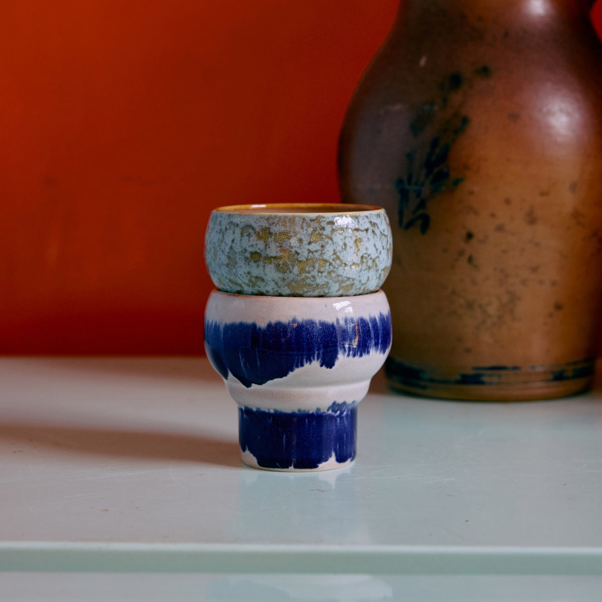 Twee gestapelde HK Living 70s Ceramics Lungo Mokken Symbolist kopjes staan op een lichtblauw oppervlak. Het onderste kopje heeft vette blauwe penseelstreken, het bovenste heeft een gespikkeld blauwgroen glazuur. Een bruine vaas verschijnt op de achtergrond tegen een oranje muur.