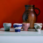 Twee kleine, uniek geglazuurde HK Living 70s Ceramics Lungo Mokken Expression kopjes van HK Living staan voor een grote bruine kan met handvat, afgezet tegen oranje en uitgestald op een bleek oppervlak.