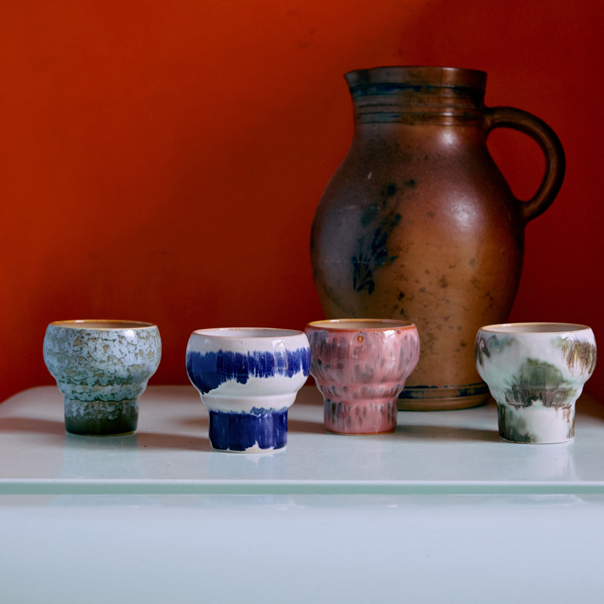 Twee kleine, uniek geglazuurde HK Living 70s Ceramics Lungo Mokken Expression kopjes van HK Living staan voor een grote bruine kan met handvat, afgezet tegen oranje en uitgestald op een bleek oppervlak.