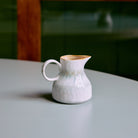 Het HK Living 70s Ceramics Melkkannetje Petal, een klein wit kannetje van keramiek met een handvat, staat op een gladde lichtgrijze tafel. De zacht vervaagde groene en bruine achtergrond benadrukt de charme van dit handgemaakte servies.