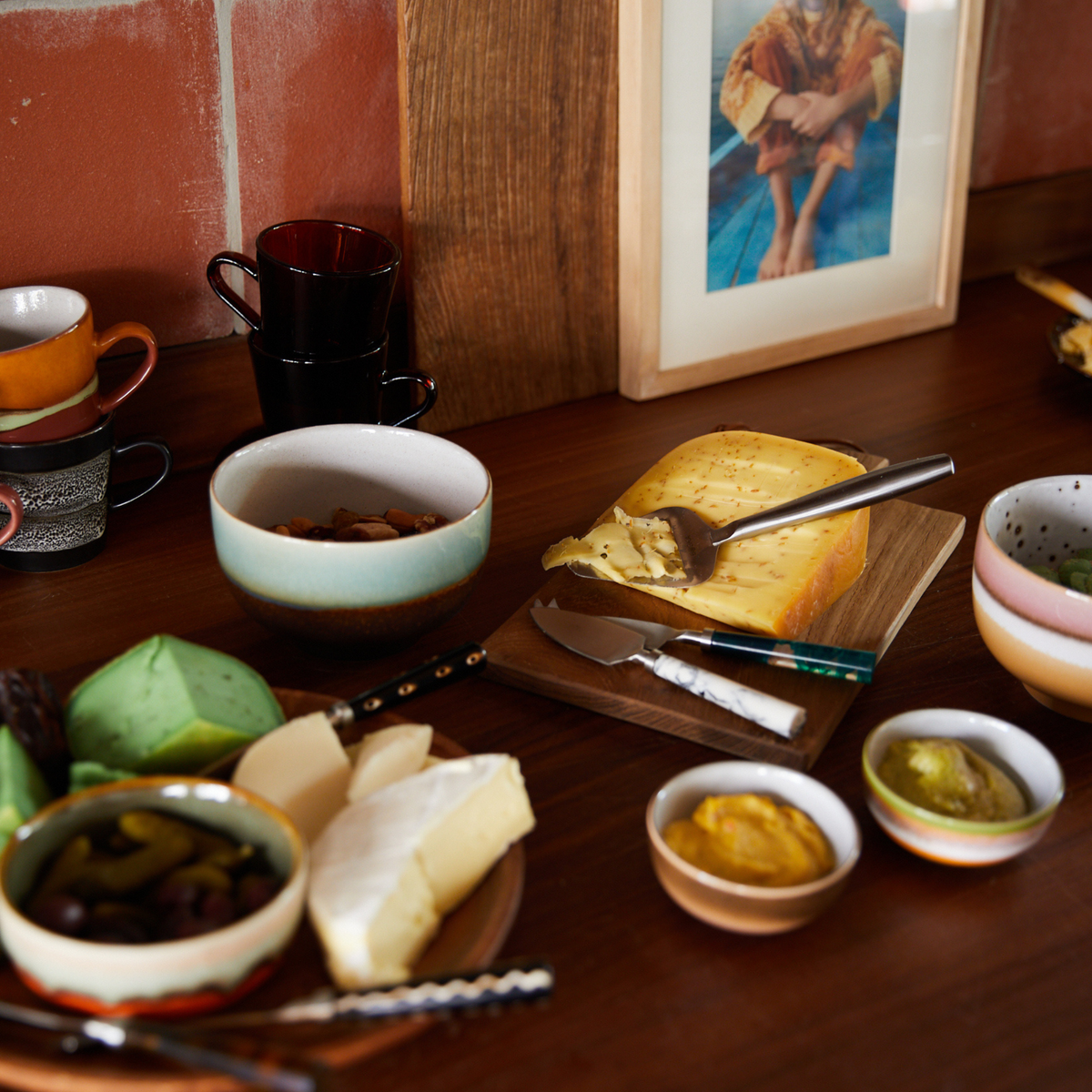 Een houten tafel gedekt met kazen, schaaltjes met noten, olijven, smeersels en kopjes, schoteltjes en de Noodle Kom Depths D13 van HK Living 70s Ceramics. Tegen de bakstenen muur staat een ingelijste foto; messen en lepels liggen naast de kazen en de keramische kom.